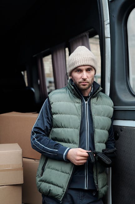 A man wearing a light grey knit beanie, a dark navy jacket with blue accents, and a green padded vest standing inside the back of a moving van. He is holding a black strap used for securing furniture