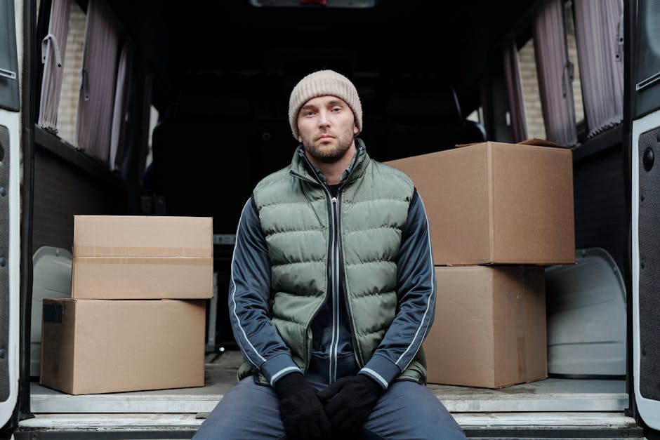 A man wearing a light grey knit beanie, a dark navy jacket with blue accents, and a green padded vest standing inside the back of a moving van. He is holding a black strap used for securing furniture