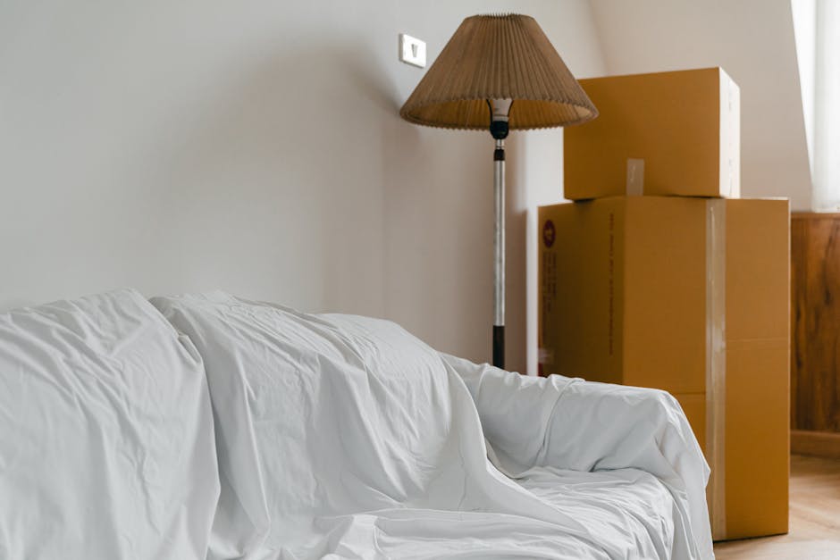 Inside a residential living space showing a white sofa covered with a white sheet on the left side of the image. Behind the sofa, there is a tall, beige floor lamp with a lampshade turned slightly to 