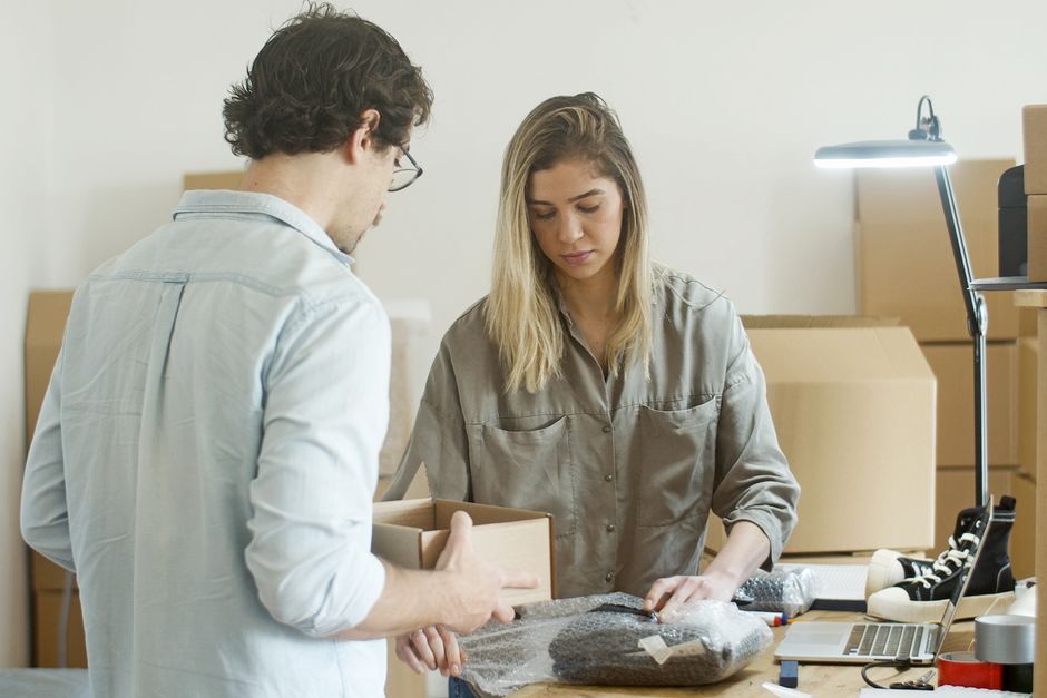 Two individuals are engaged in packing and preparing items for a furniture transport or home relocation in a well-lit indoor environment. The man, wearing glasses and a light blue shirt, holds an empt