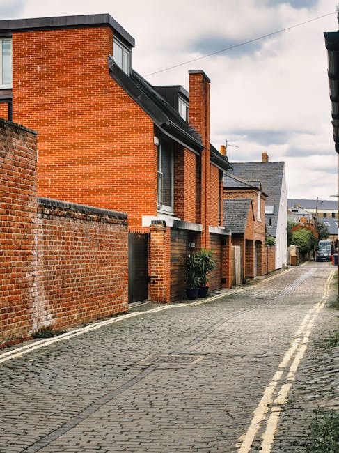A cobbled residential street with brick terraced houses featuring a mix of red and grey brickwork, with some houses having small front gardens, and others with paved entrances. The buildings are two t