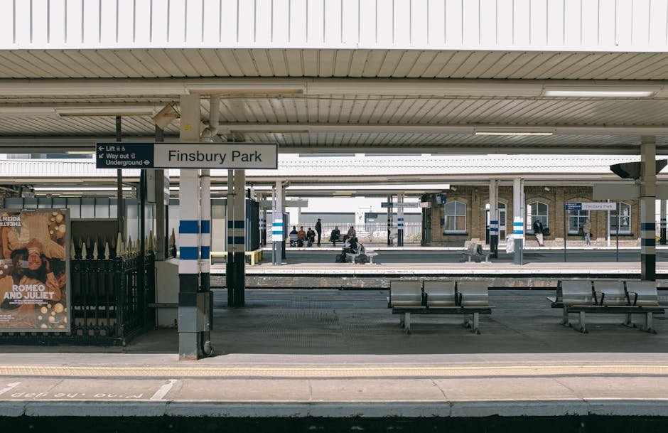 The image depicts a view of Finsbury Park railway station platform with a white and blue sign displaying the station name. The platform is covered by a metal roof with fluorescent lighting, supported